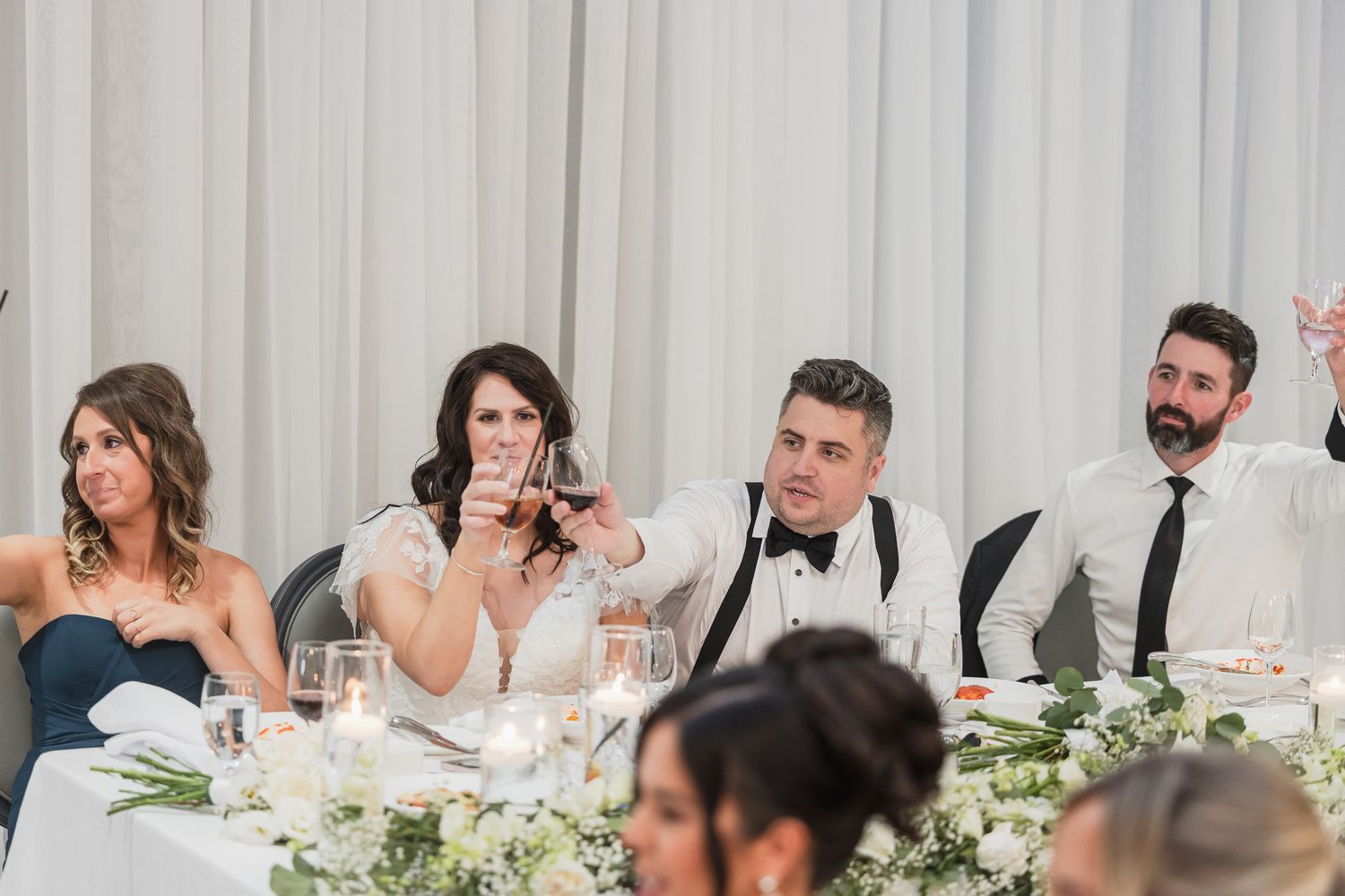 Wedding party raises glasses in a toast at the head table, decorated with white flowers and candles.