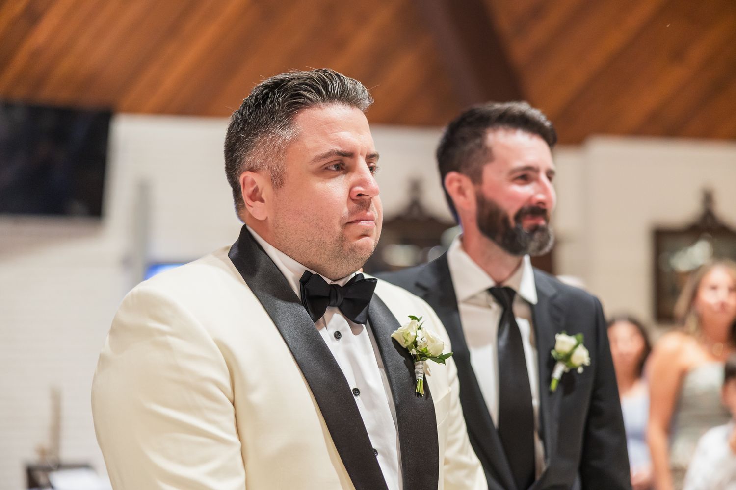 Emotional groom in white tuxedo stands at altar with groomsman beside him during wedding ceremony.