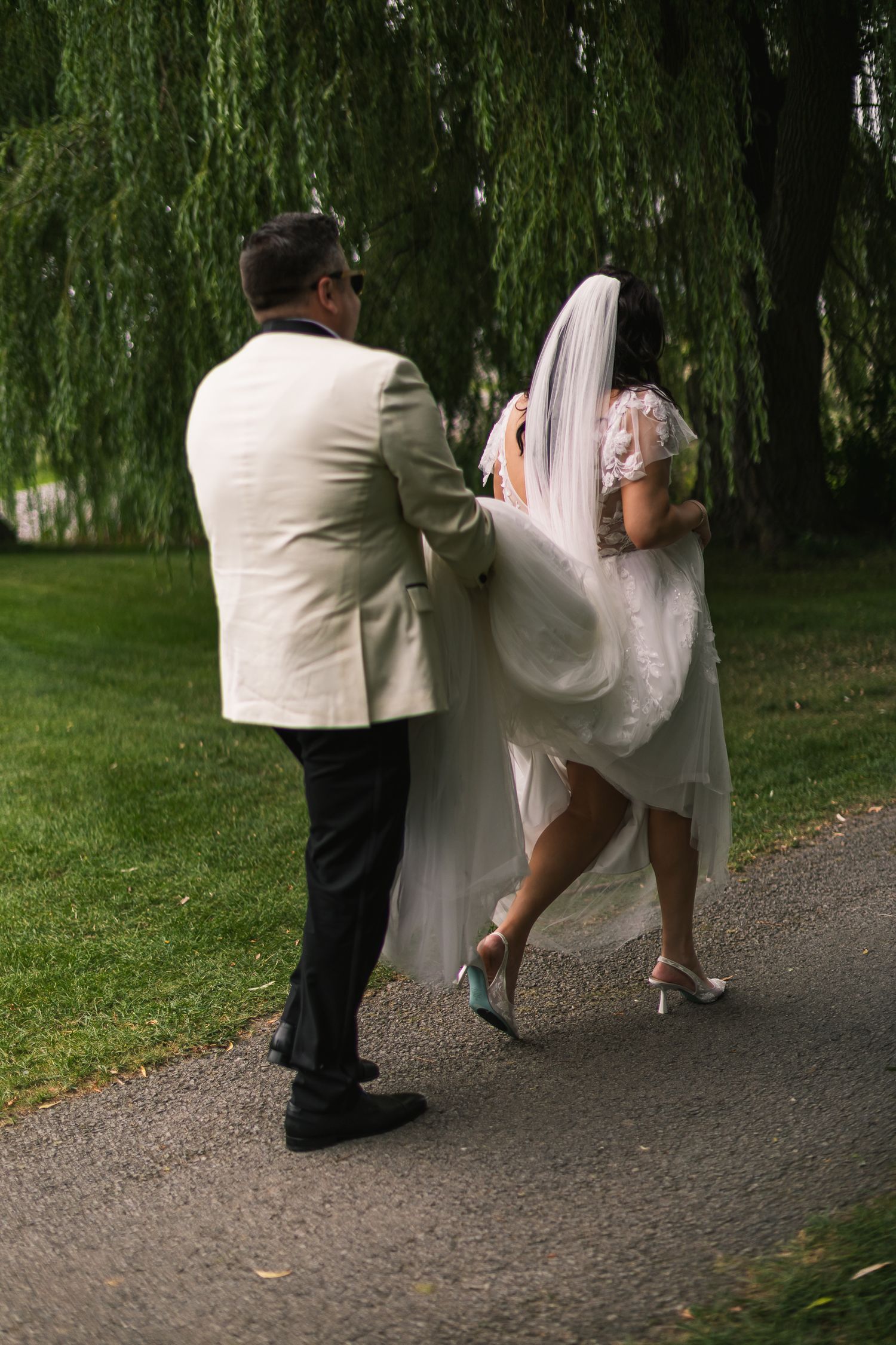Bride and groom walking hand in hand on a garden path beneath a weeping willow tree.