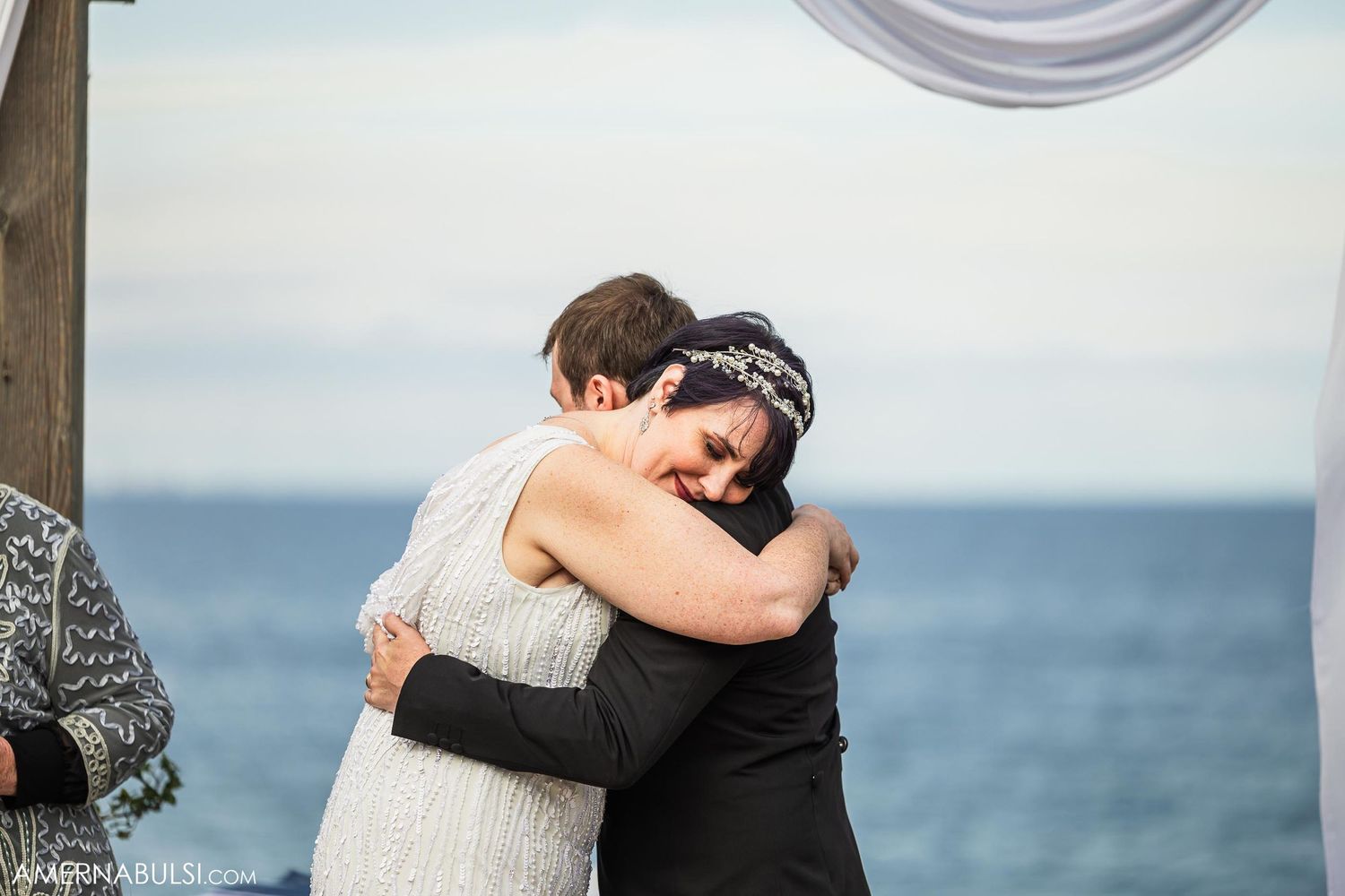 Emotional embrace during seaside wedding ceremony under white draped arch.