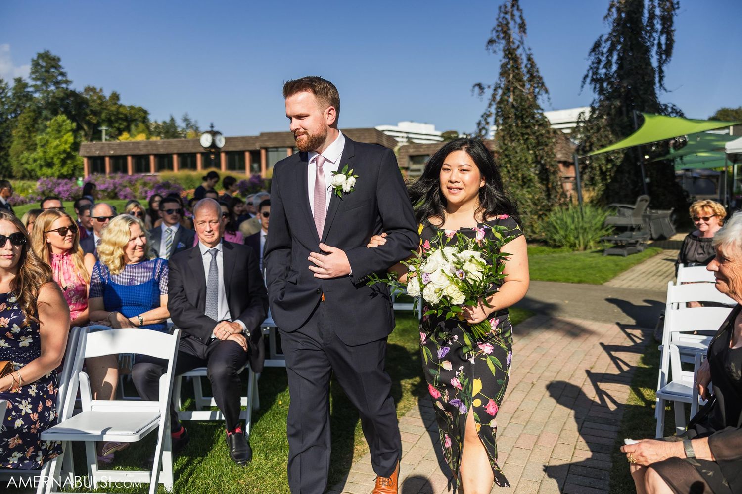 Wedding party walking down outdoor aisle with seated guests and green shade sail overhead.