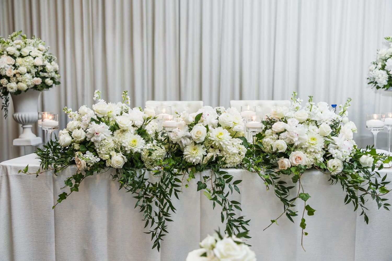 Elegant white floral arrangement with greenery decorates wedding reception head table.