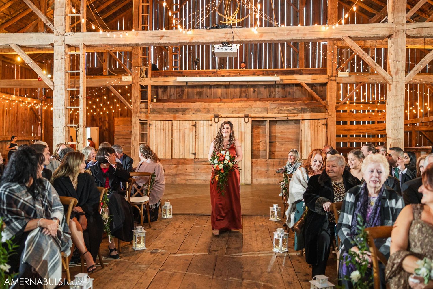 Bridal party member in burgundy dress walks down aisle of rustic barn wedding ceremony with string lights overhead.