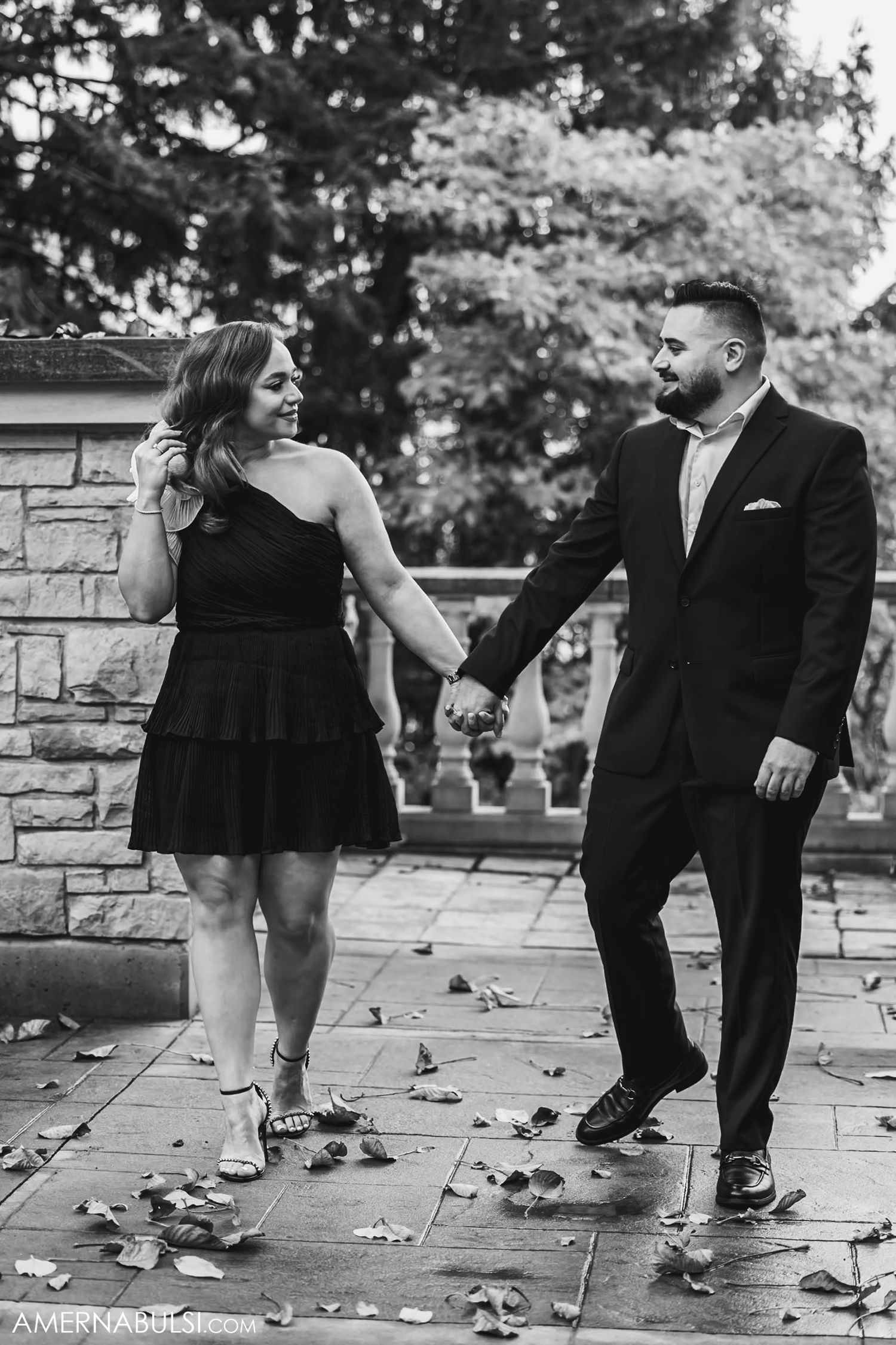 Couple in formal wear holding hands and walking on stone patio covered in autumn leaves.