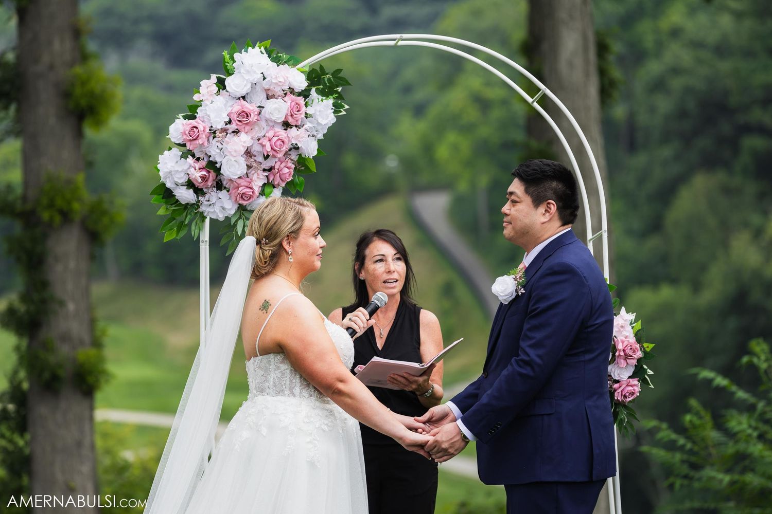 Wedding ceremony taking place beneath an elegant arch adorned with pink and white flowers.