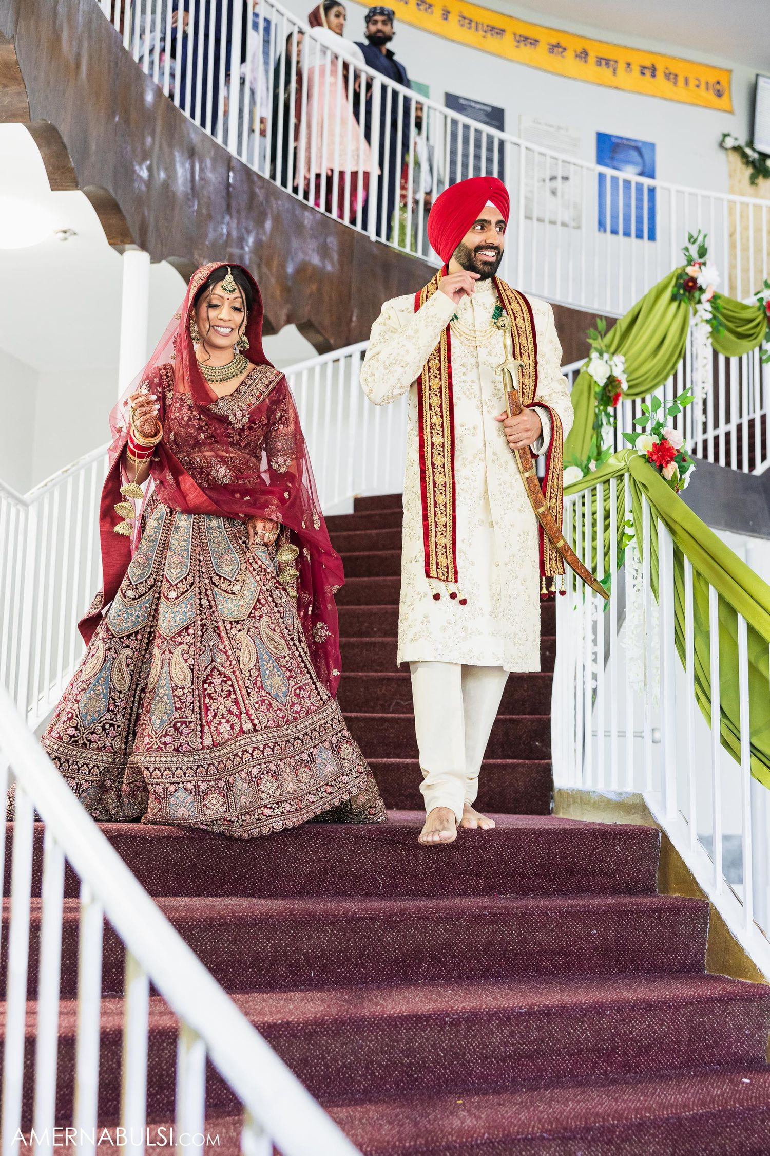 Couple in traditional Sikh wedding attire descending decorated staircase with red carpet.