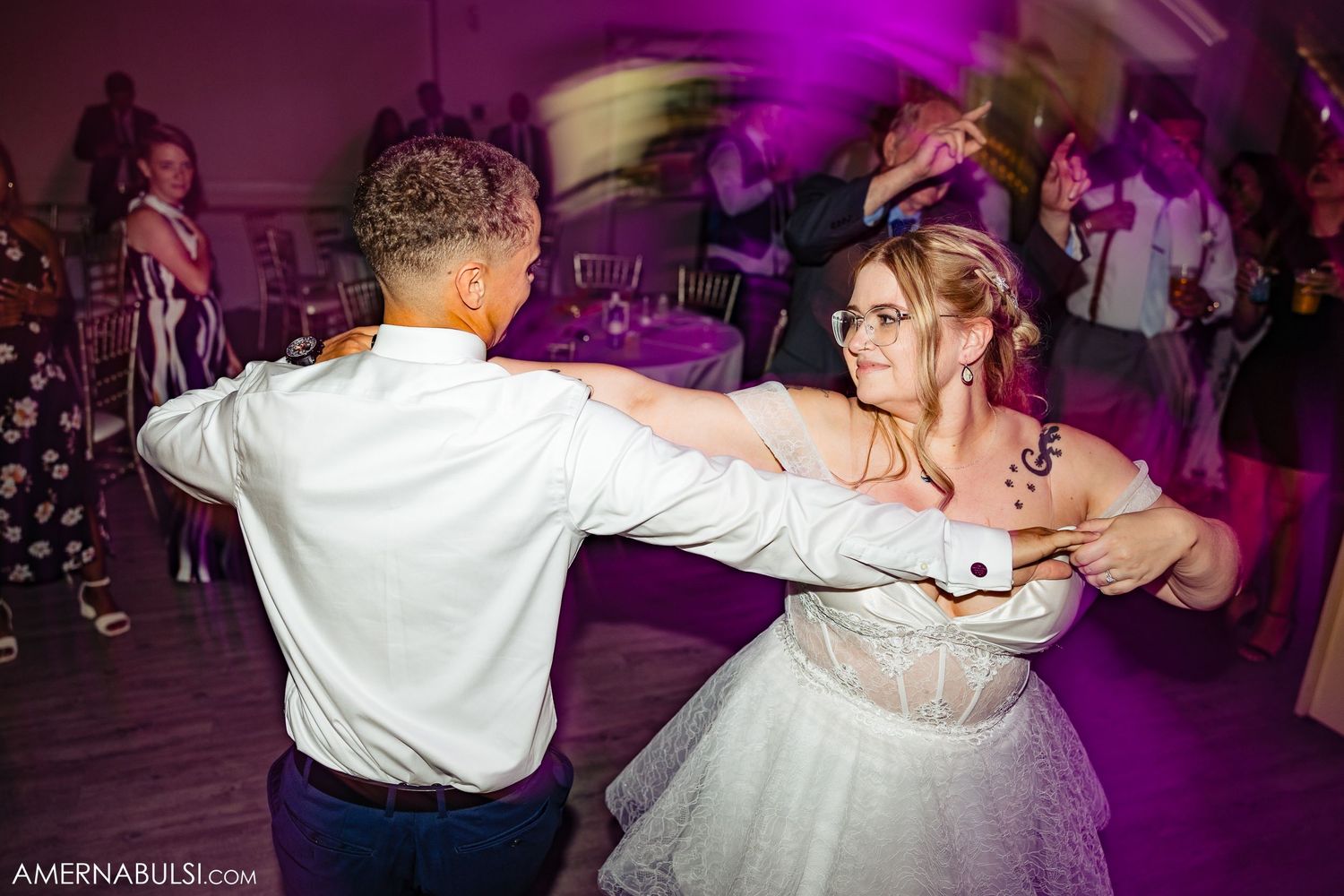 A couple dances under dramatic purple lighting at a wedding reception with other guests visible in the background.