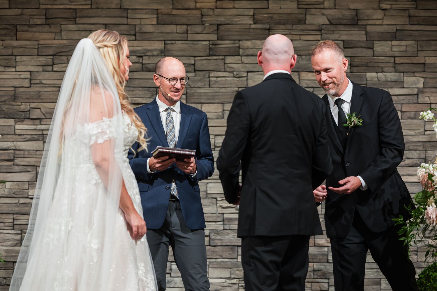 Outdoor wedding ceremony takes place against stone wall backdrop with officiant and wedding party.