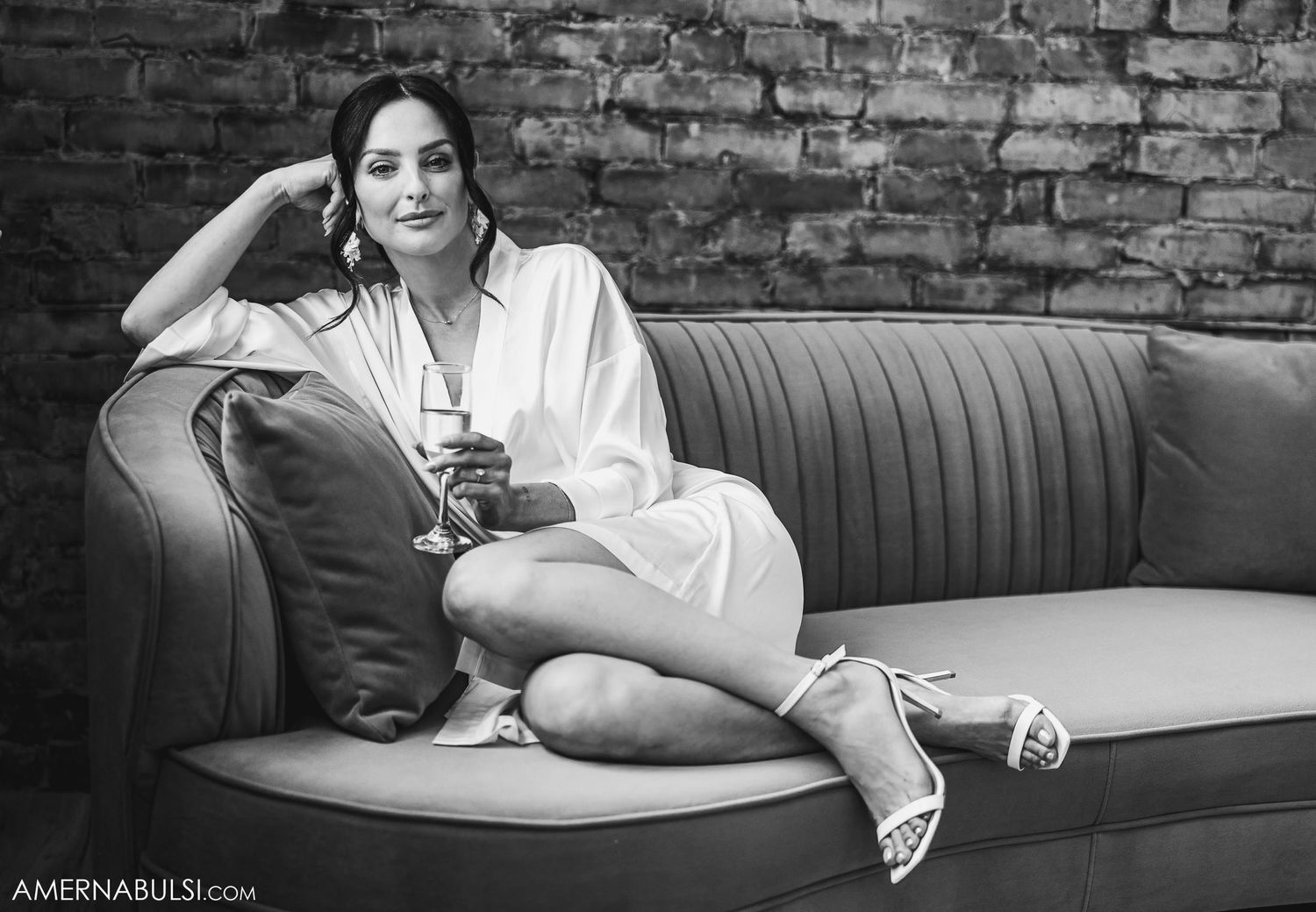 Woman in white robe adjusts wedding dress hanging on exposed brick wall.