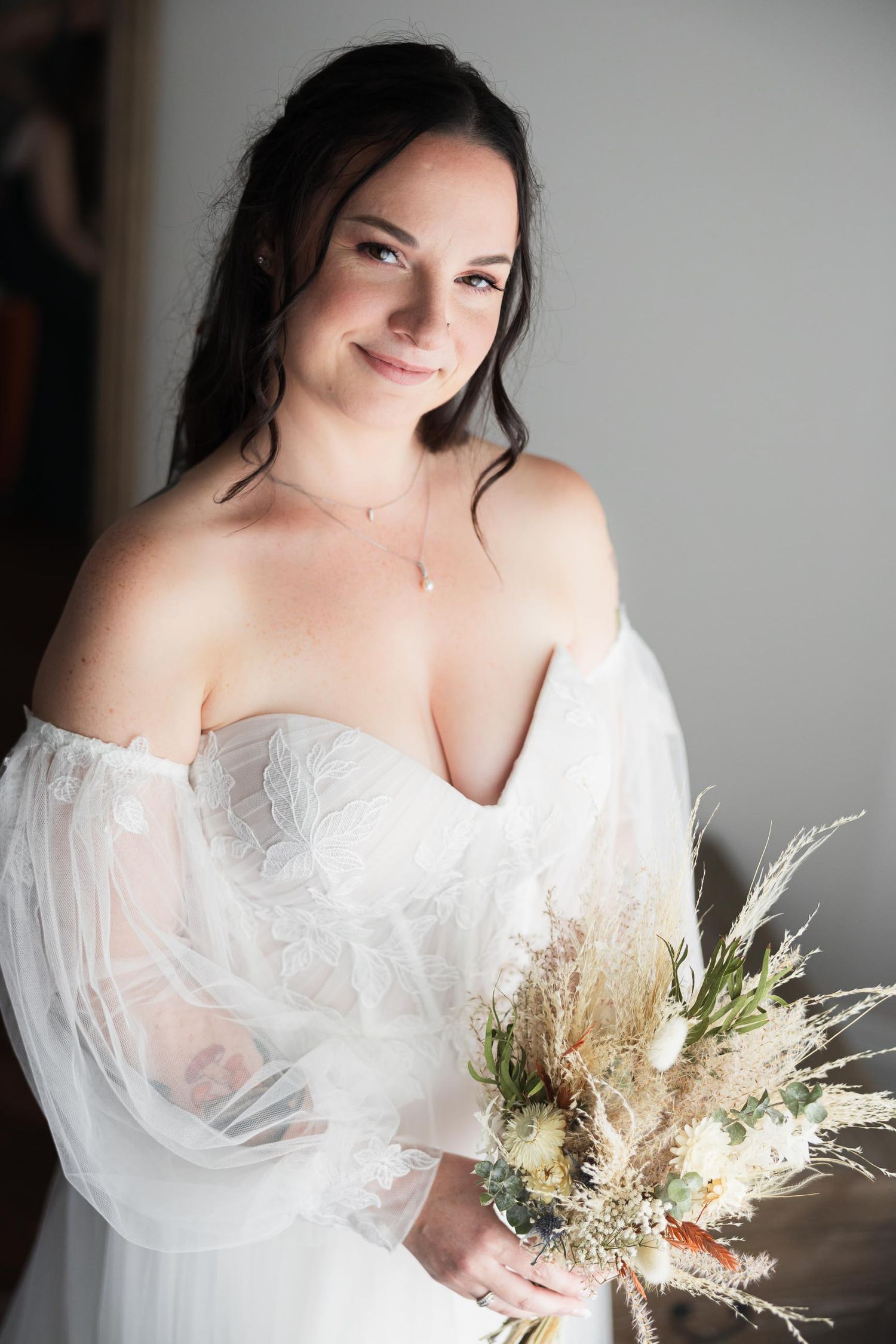 A bridal portrait showing an off-shoulder white dress and dried floral bouquet against a gray background.