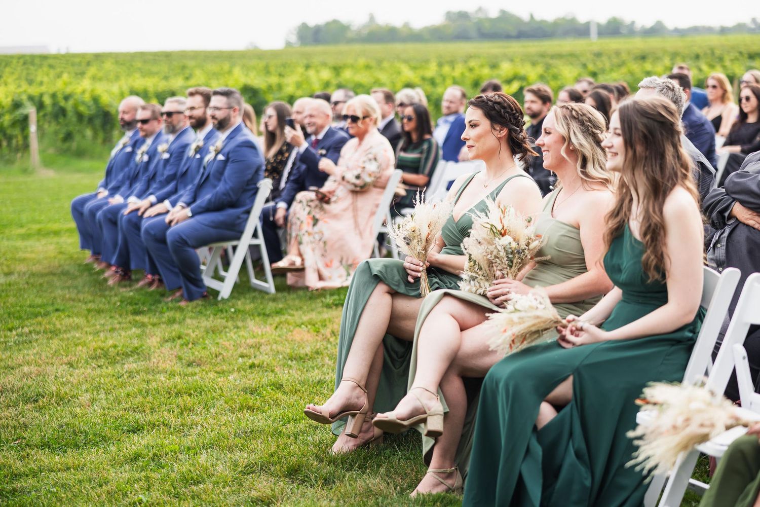 Wedding guests seated in rows of white chairs with attendants in navy and emerald green at a vineyard venue.