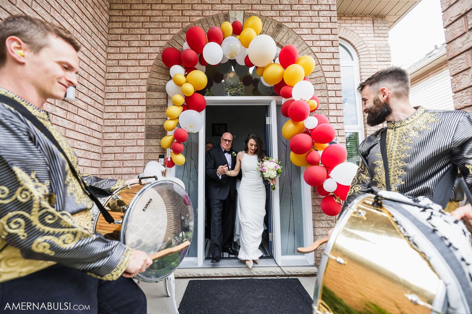 Couple emerges from doorway under colorful balloon arch with drummers in foreground.