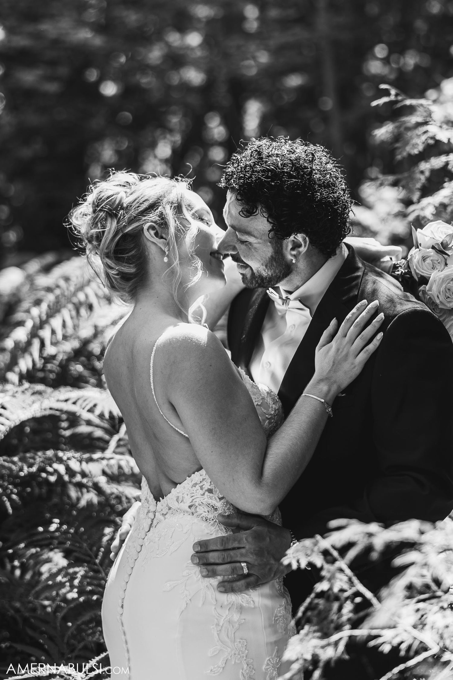 Black and white portrait of couple sharing intimate moment among ferns in dramatic lighting.