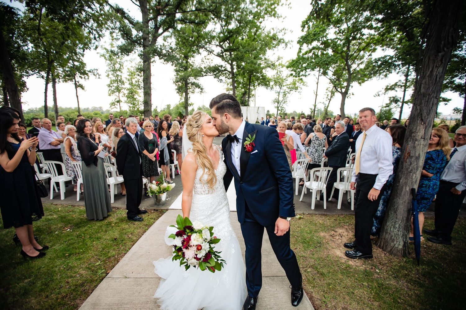 Guests gather outdoors at a wedding ceremony under tall trees on a sunny summer day.