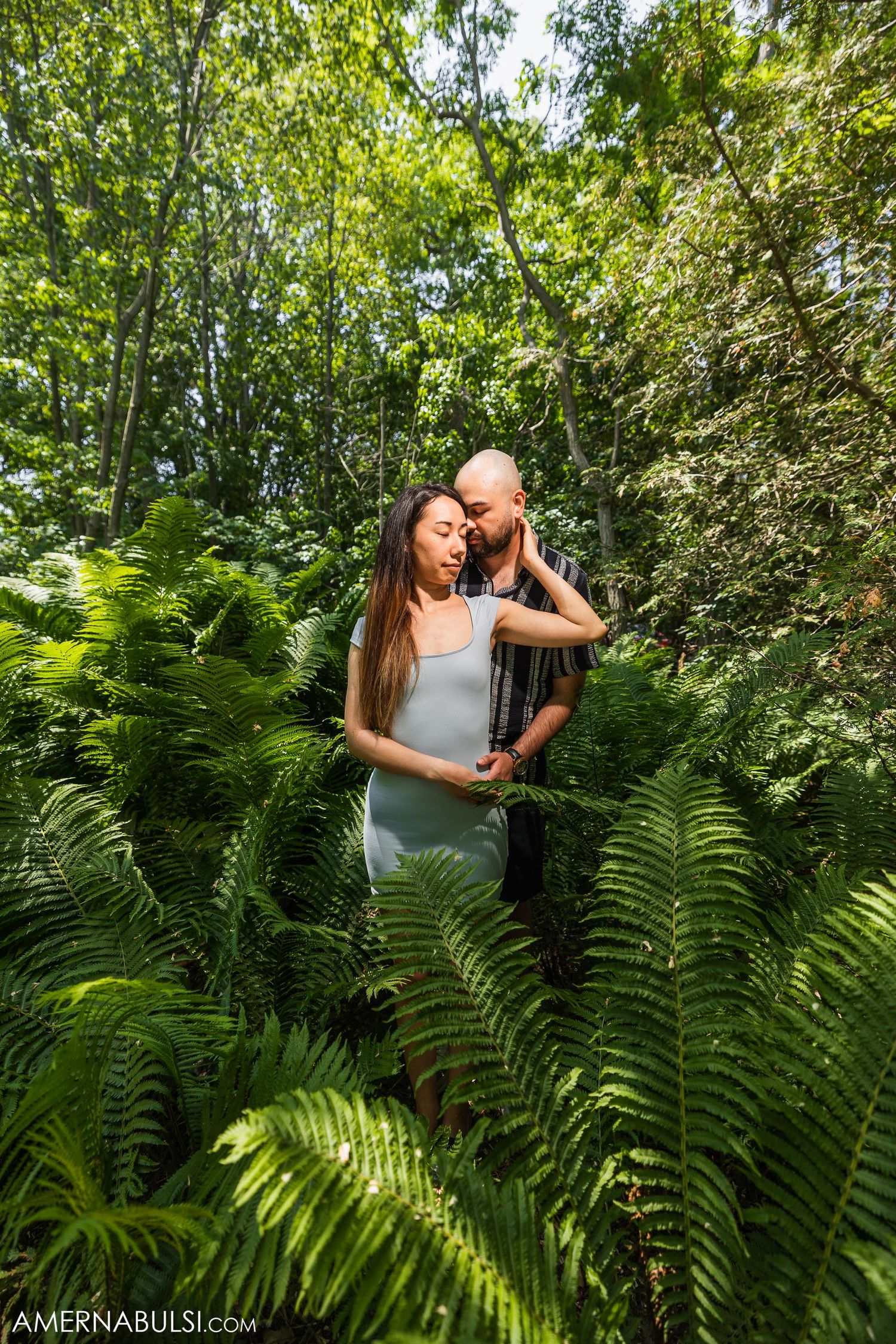 A couple shares an intimate embrace among wild ferns in dappled sunlight.