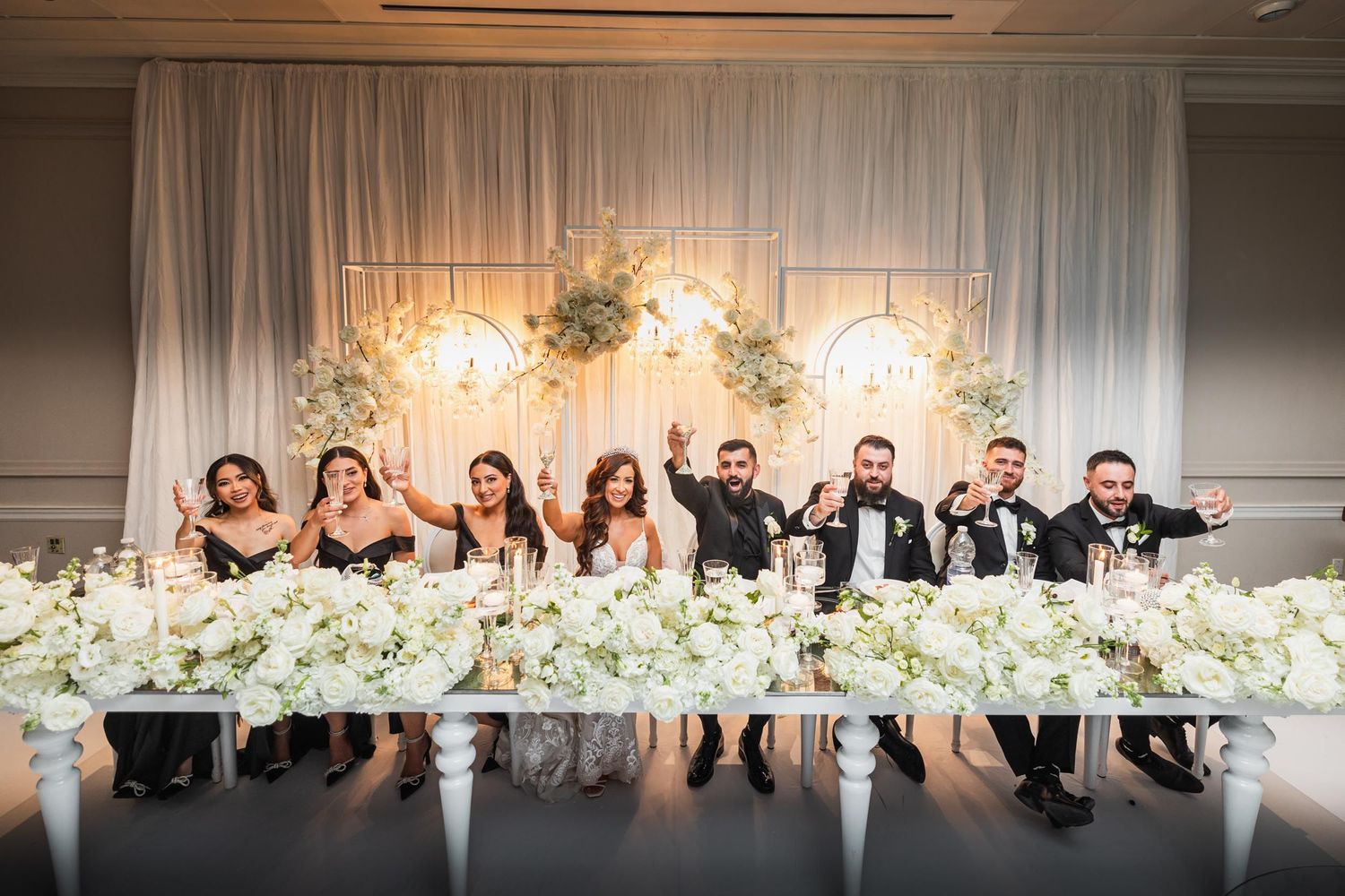 Wedding party seated at elegant head table decorated with white floral arrangements and draped backdrop.