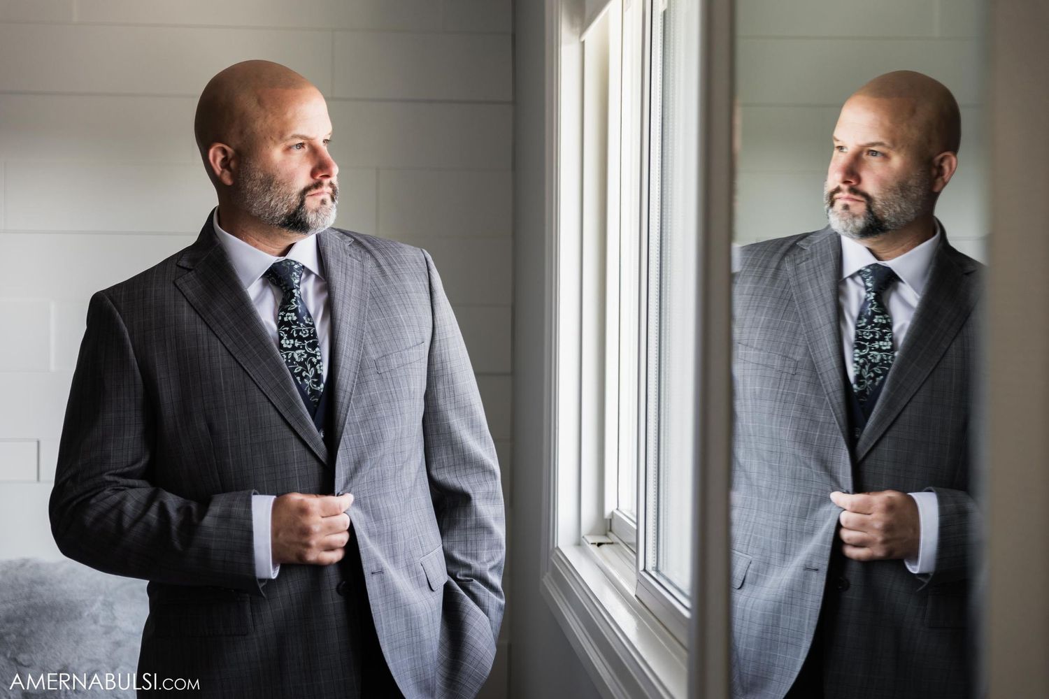 Reflection of person in grey suit adjusting tie by window.