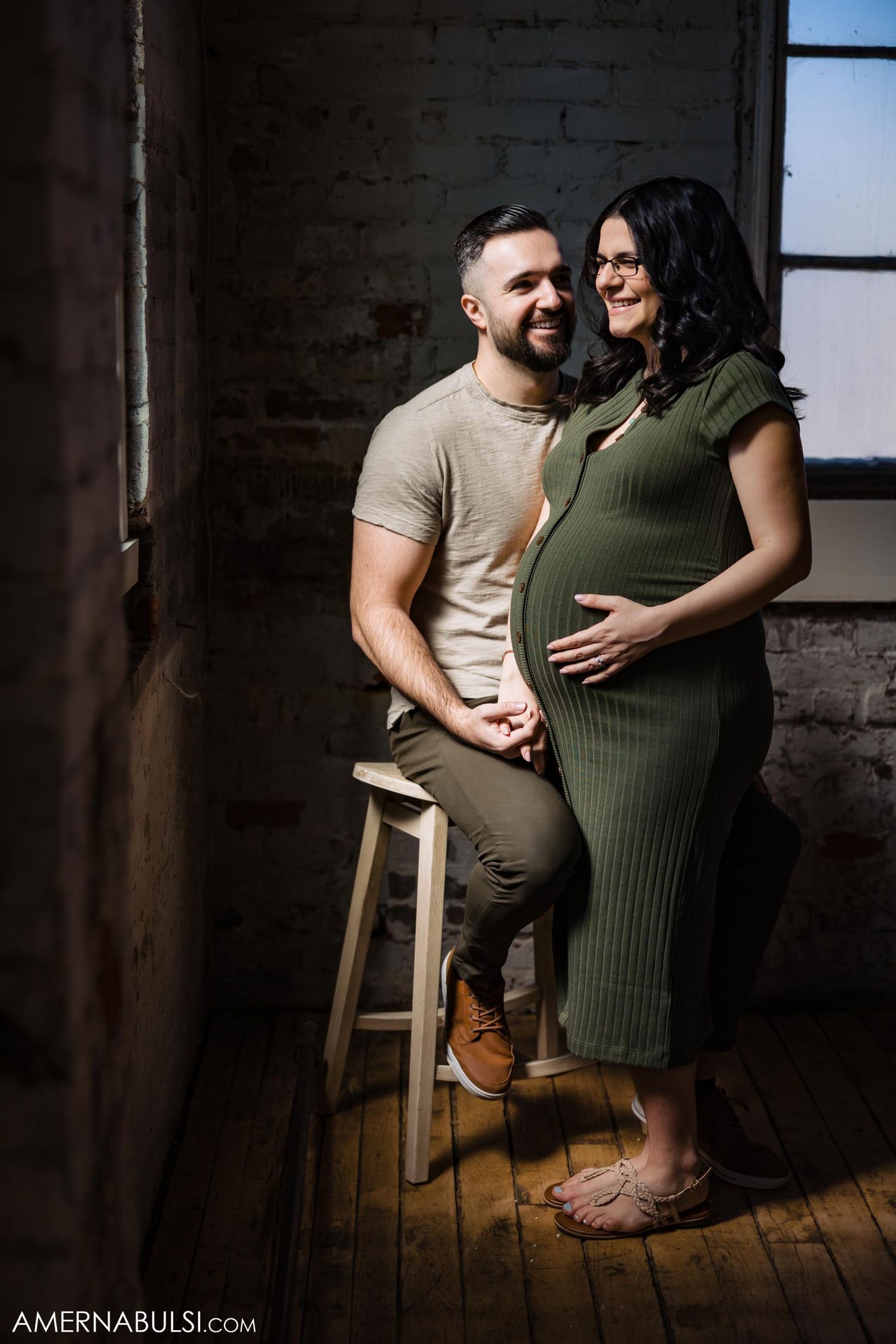 Industrial maternity photoshoot with couple posing by wooden stool against brick wall.