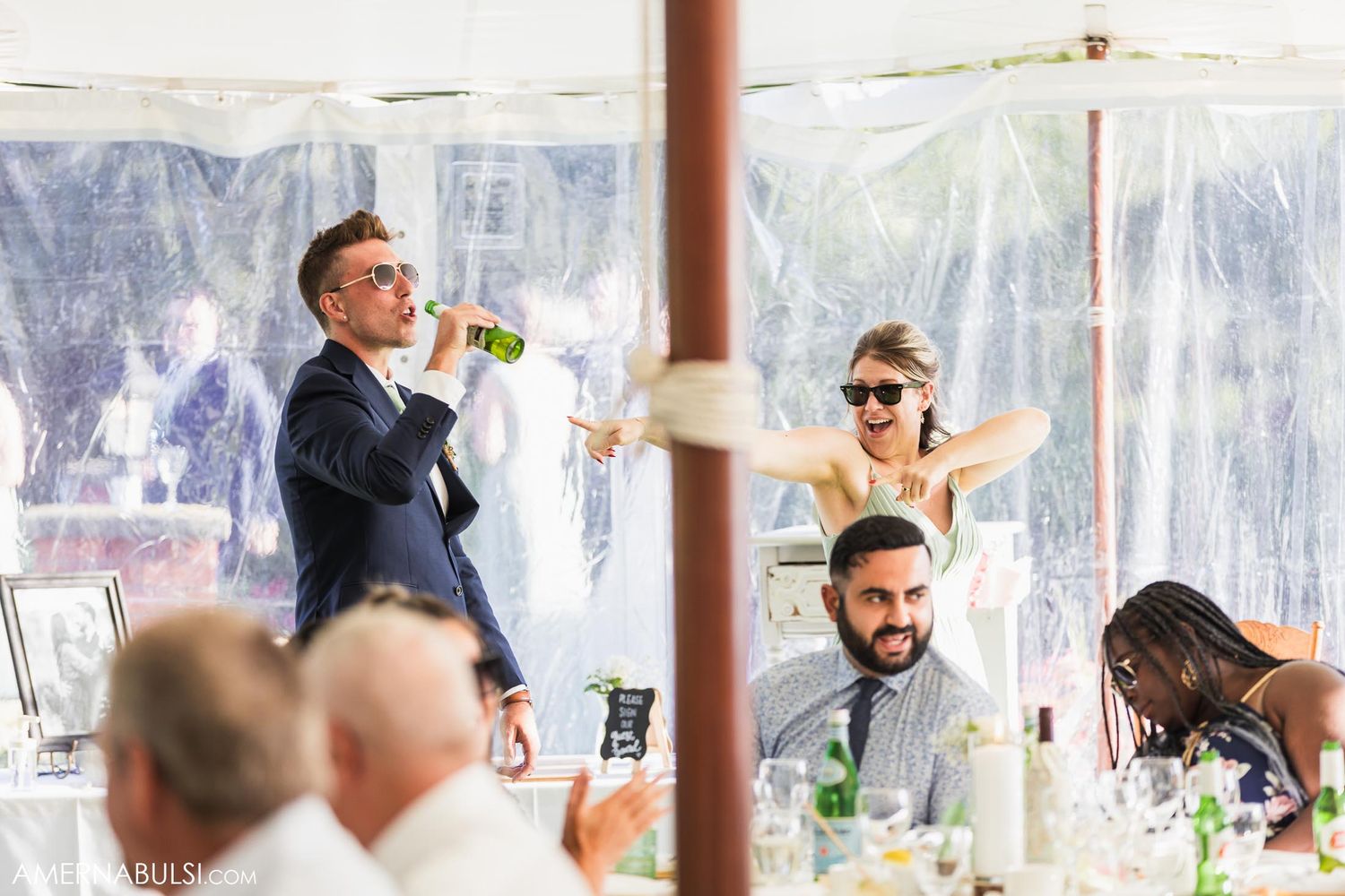 Lively wedding reception scene with guests celebrating under clear tent walls.