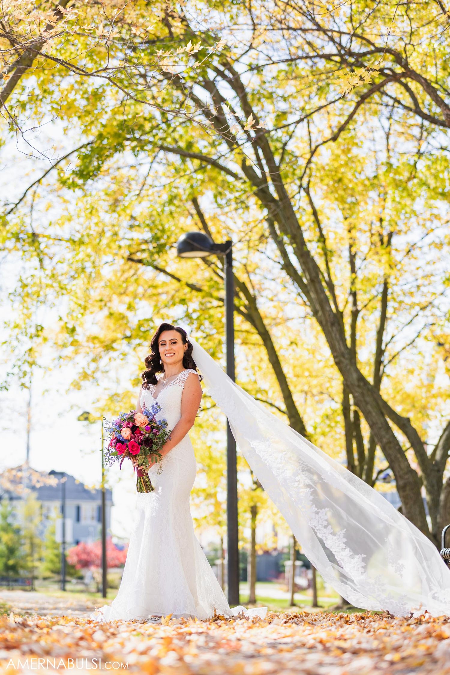 A bride with flowing veil poses in autumn foliage holding a pink and red bouquet.