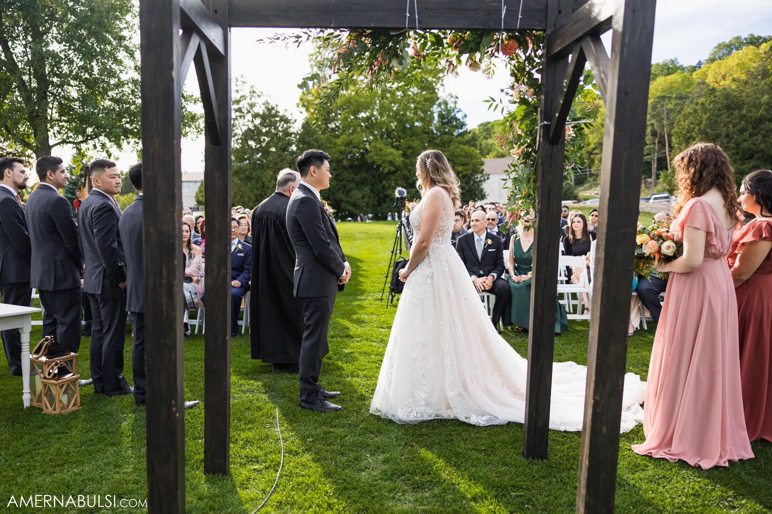 Outdoor garden wedding ceremony under a wooden pergola with guests seated on white chairs.