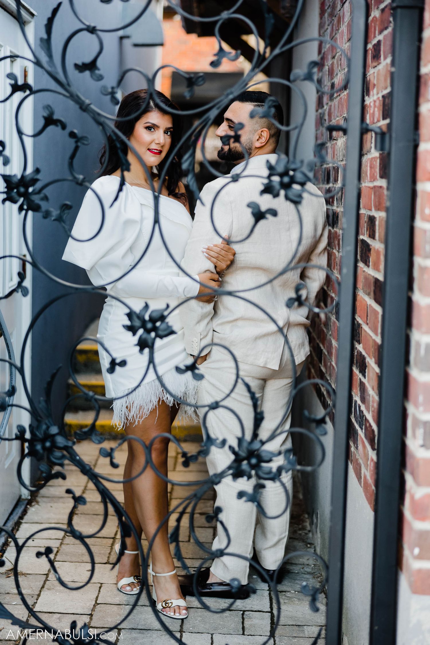 A couple in coordinated white outfits pose against decorative iron scrollwork on a brick wall.