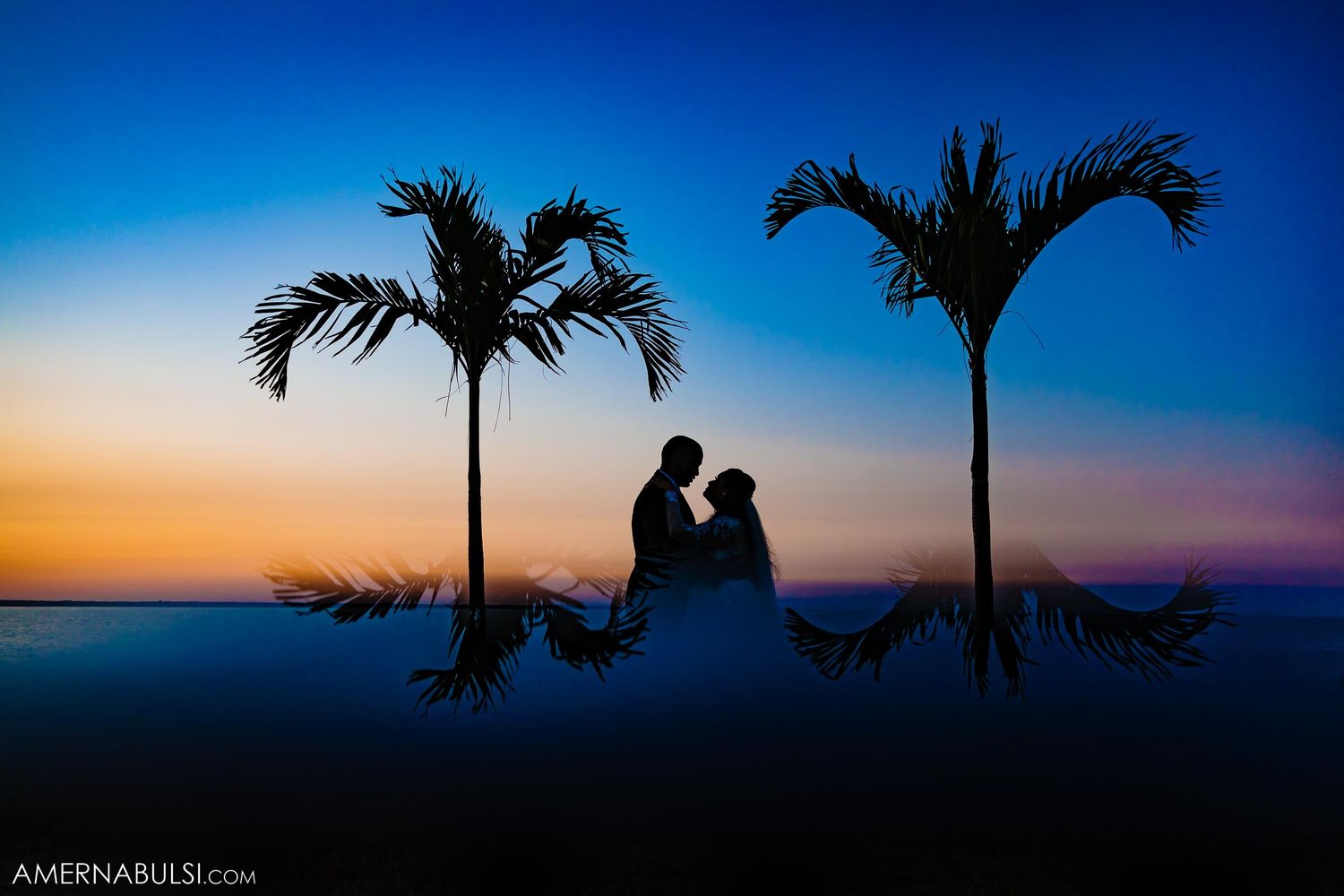 Romantic silhouette of a couple between two palm trees against a vibrant sunset and blue sky.