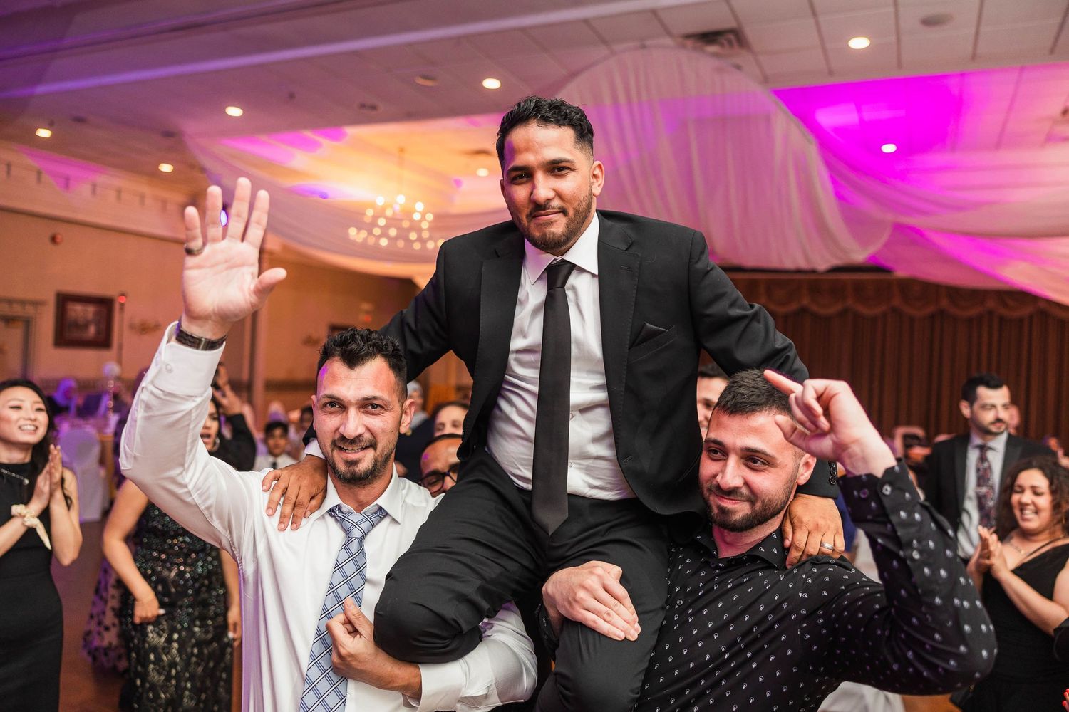 Wedding guests lifting and celebrating with the groom at a reception with pink lighting and white draping.
