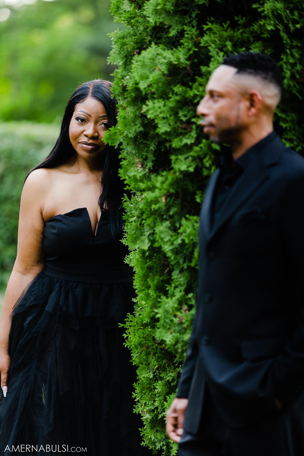 Couple in elegant black attire pose against green hedges in a garden setting.