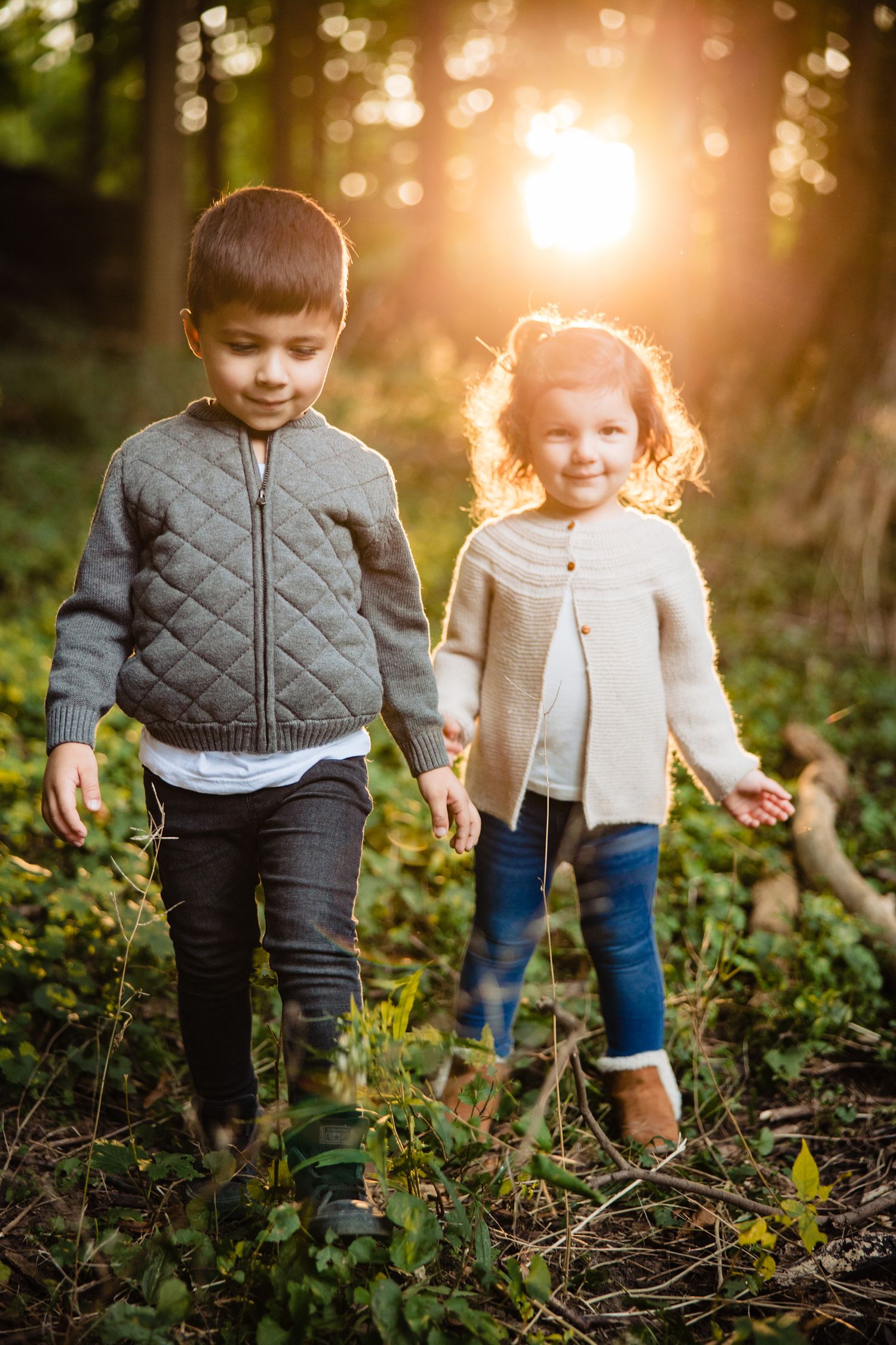 Children explore a sunlit forest path together during golden hour, wearing casual outfits and walking among fallen branches.