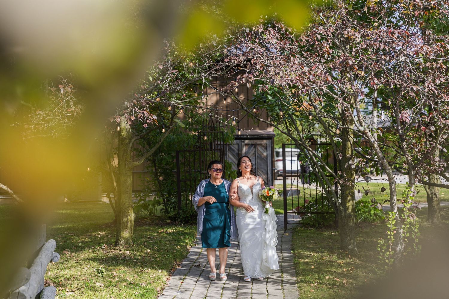 Bride walks down garden pathway with escort through blooming floral arch toward outdoor wedding ceremony.