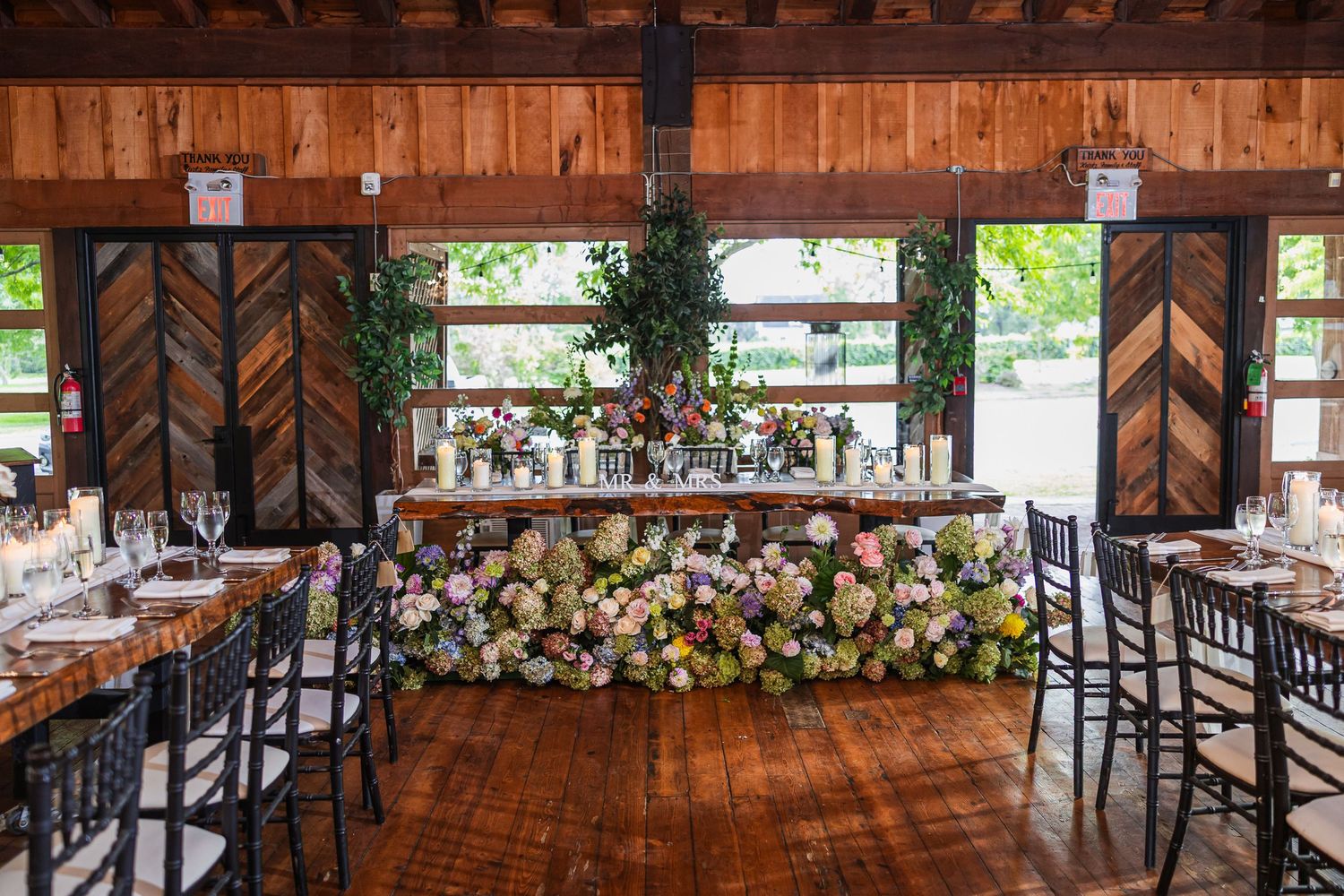 Elegant rustic wedding reception venue with floral head table, string lights, and wooden chevron doors.