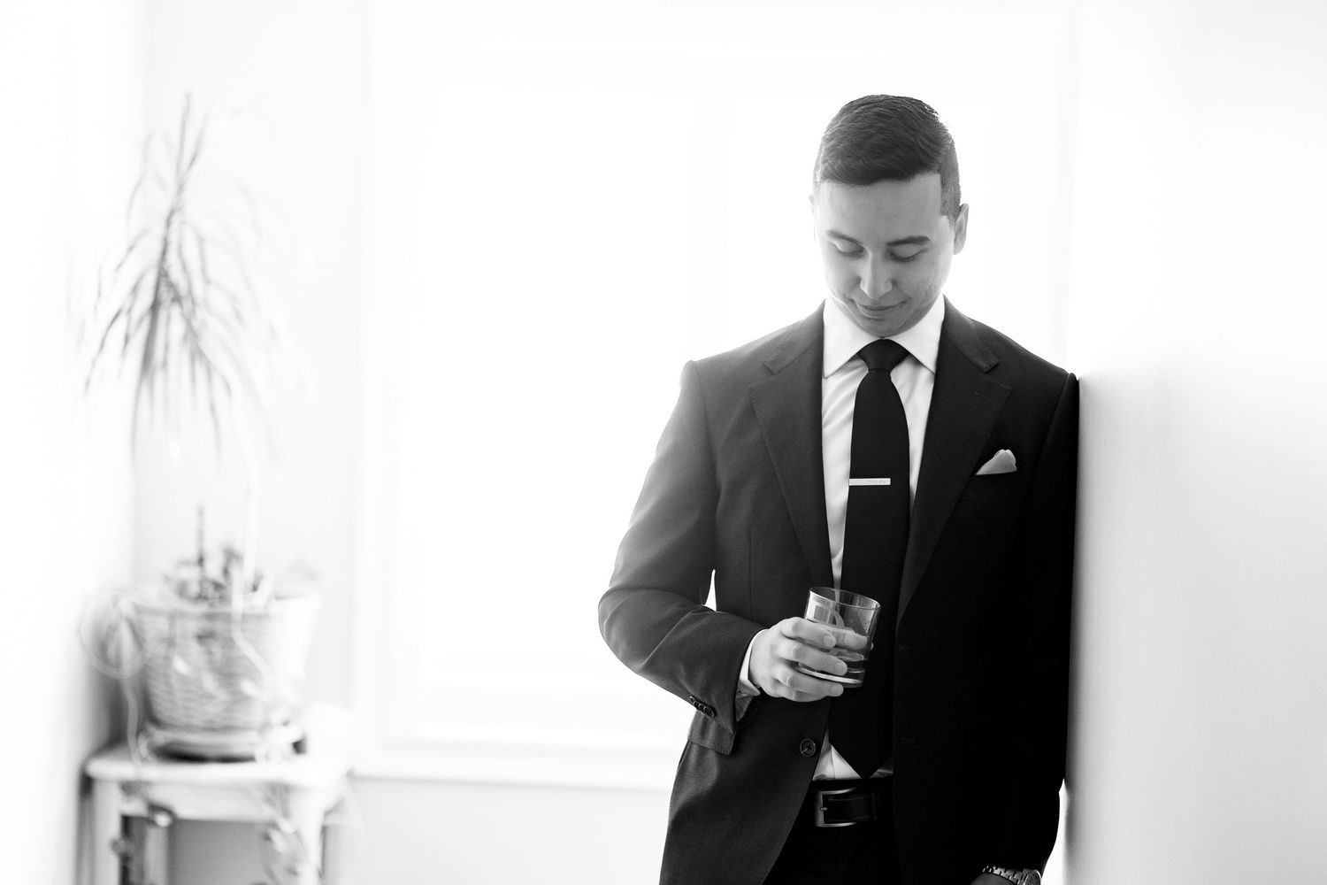 A groom in a black suit holds a glass while looking down in a bright, elegant room.