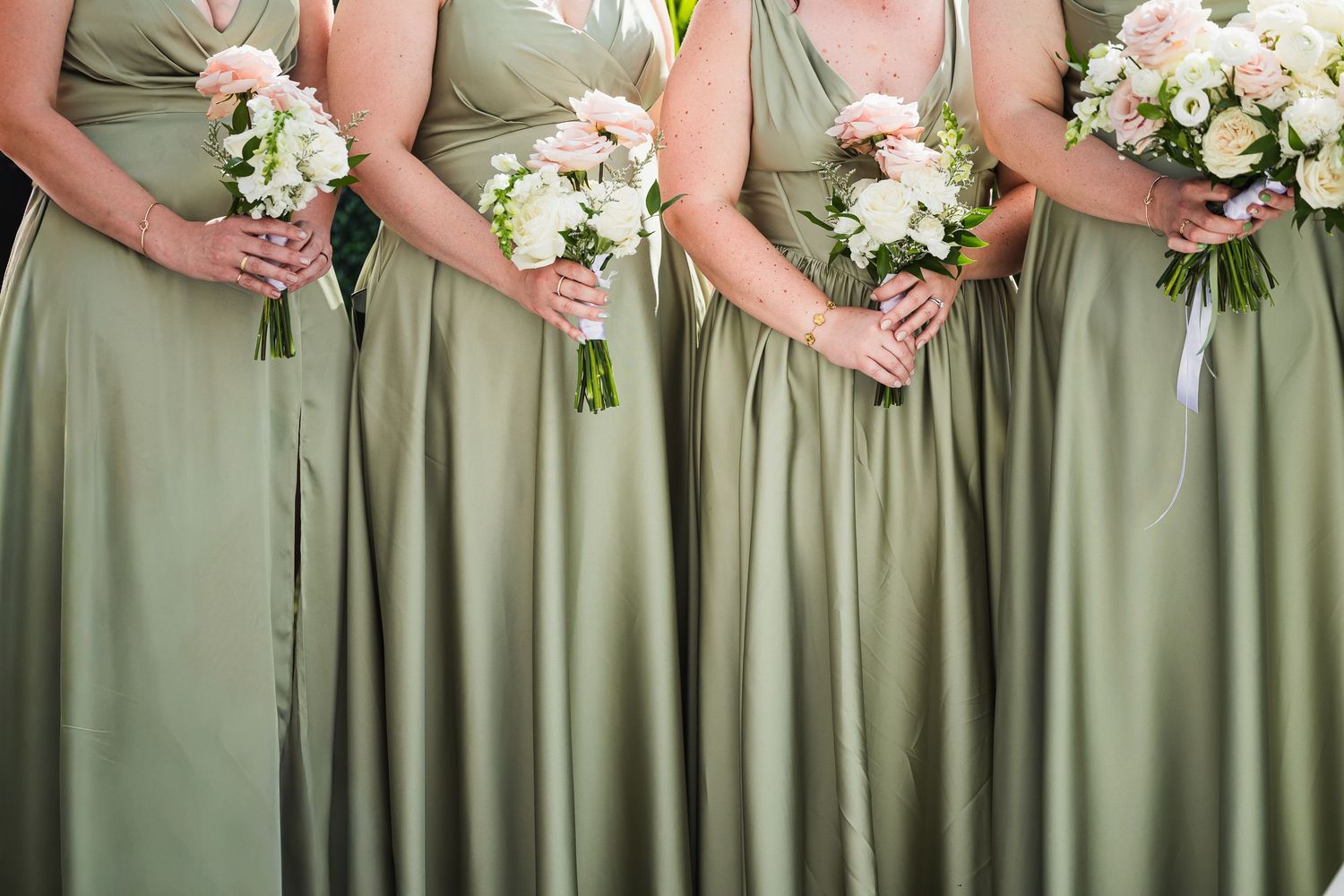 Bridesmaids in sage green gowns holding white and pink floral bouquets at wedding ceremony.