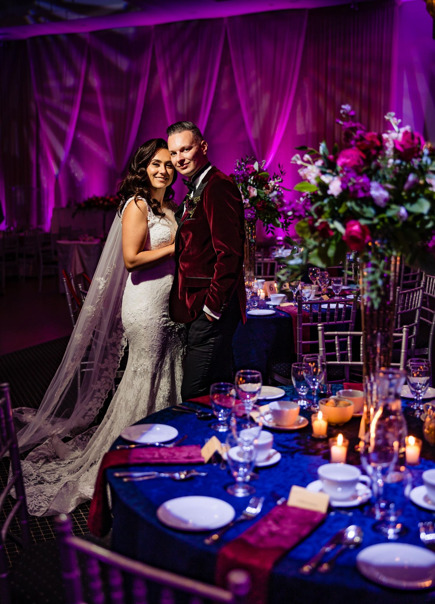 Reception tables set with blue linens and floral centerpieces under purple uplighting.