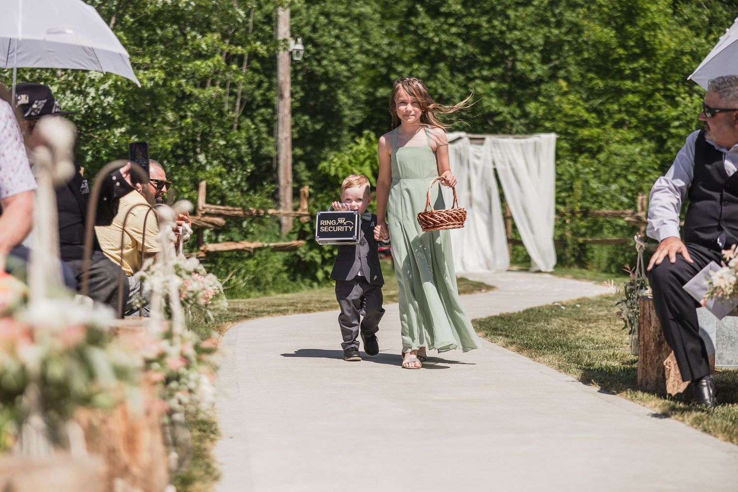 Young ring bearer and bridesmaid in sage green dress walking down outdoor wedding ceremony aisle.
