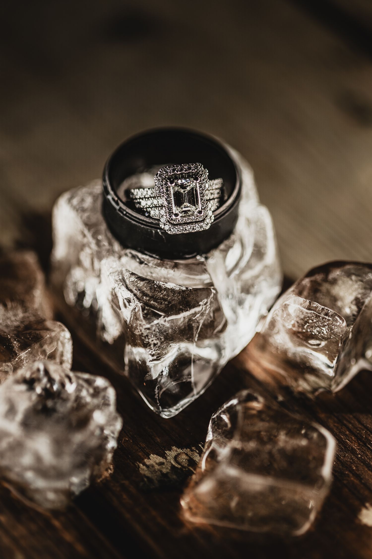 Diamond engagement ring and black wedding band resting on ice cubes on a rustic wooden surface.
