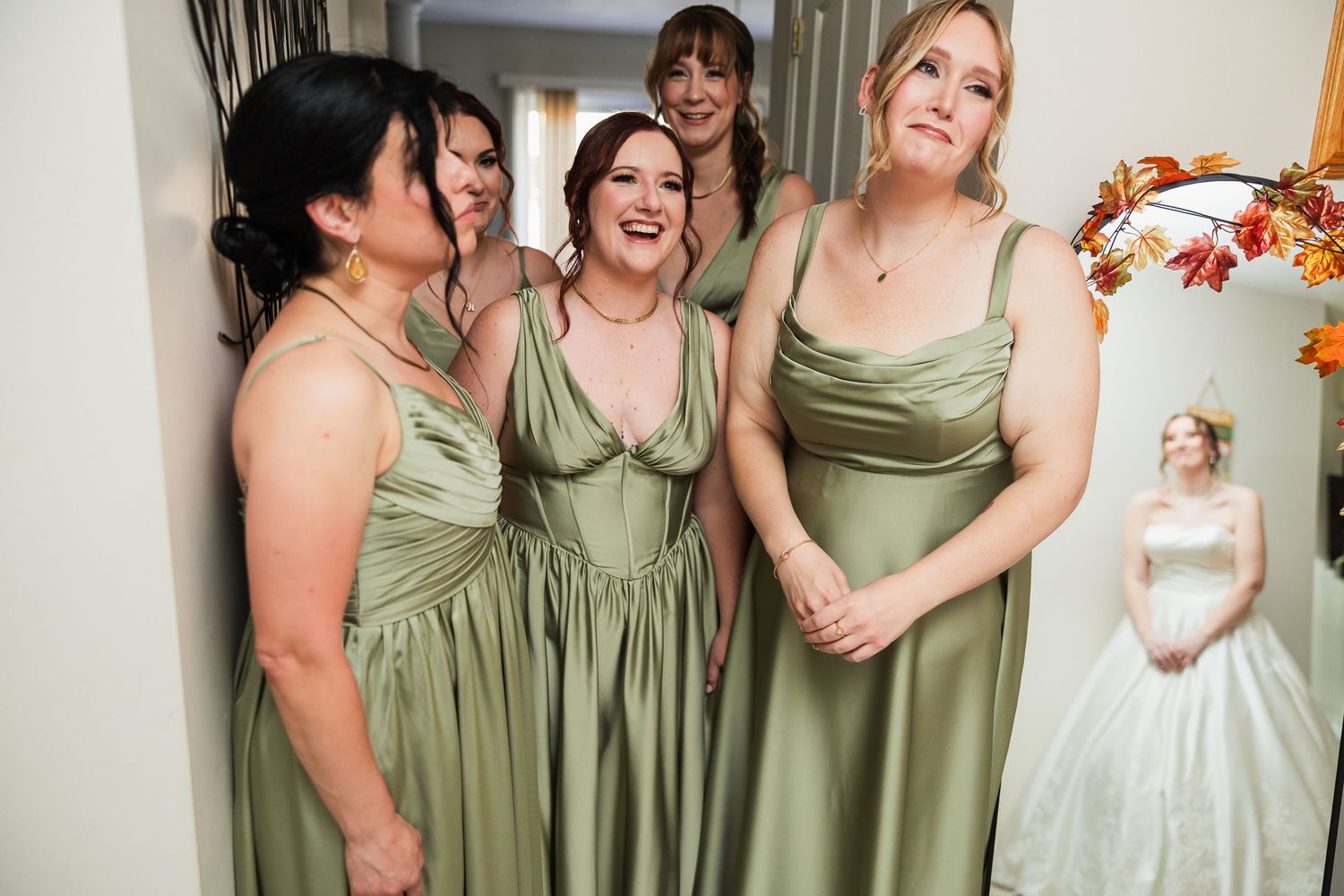 Bridesmaids in sage green dresses laugh joyfully as bride in white gown stands in background.