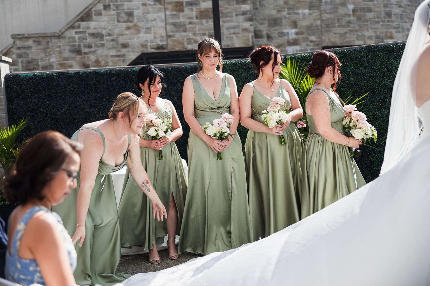 Four bridesmaids in sage green gowns holding white bouquets stand outdoors near a stone wall beside a bride in white.