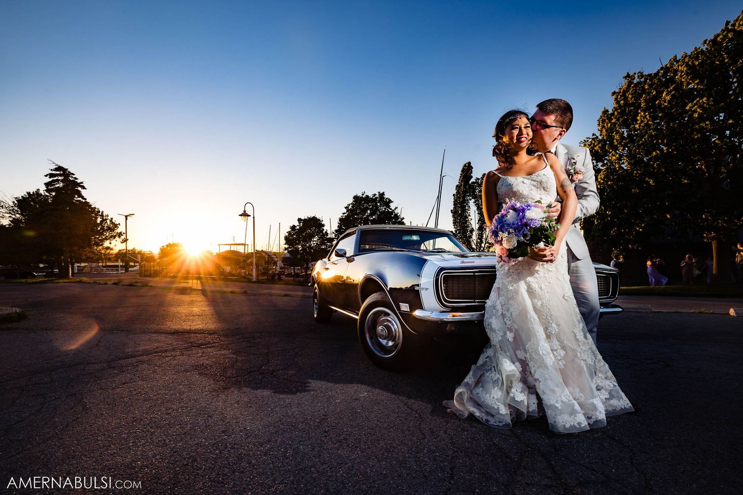 Newlyweds pose at sunset with vintage car in parking lot.