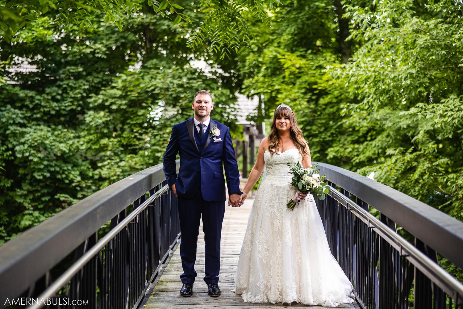 Wedding couple poses on wooden bridge surrounded by lush green forest.