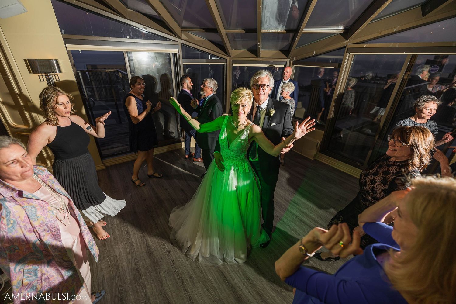 Wedding party dancing in a venue with dramatic lighting and a couple in green attire at the center.