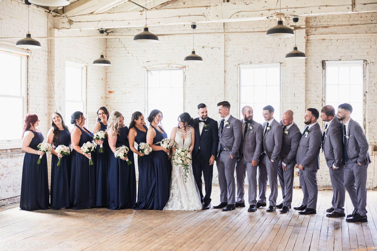 Wedding party in navy and grey attire poses in front of bright windows in industrial venue.