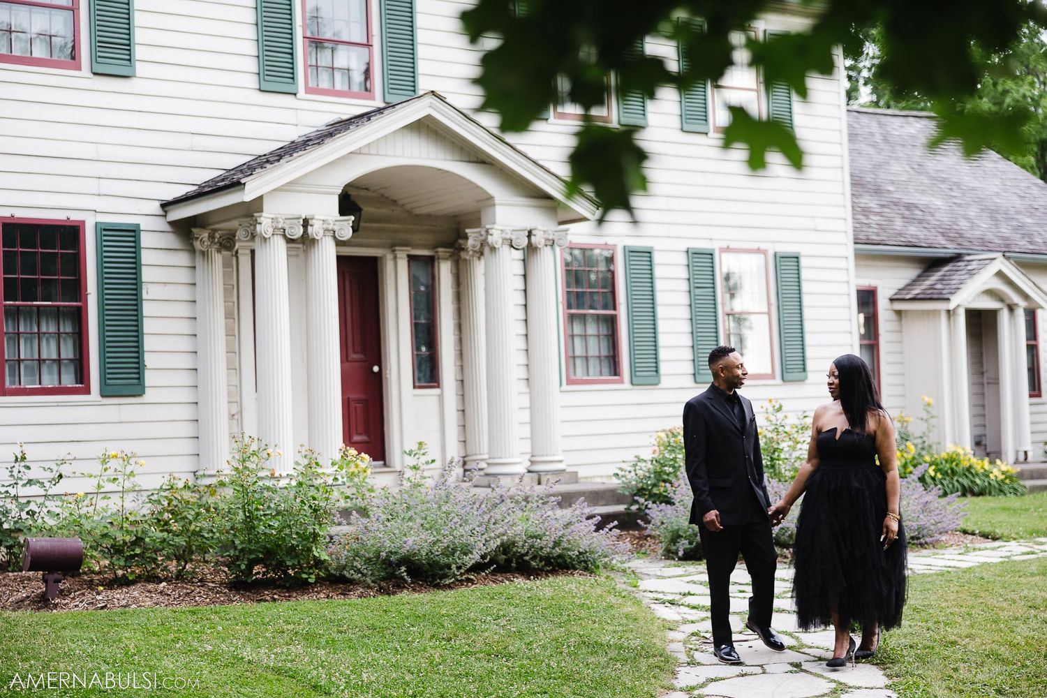 Elegant couple poses in front of a white colonial house with columns and green shutters.