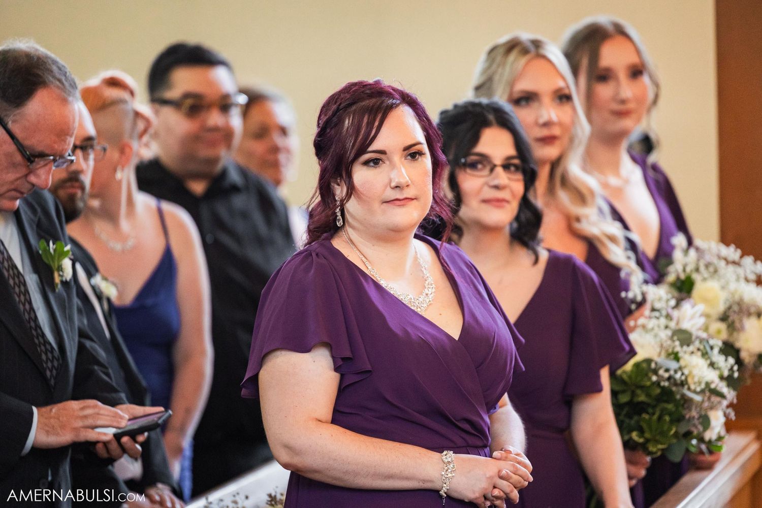 Wedding party and guests watching ceremony in church with purple bridesmaid dresses.