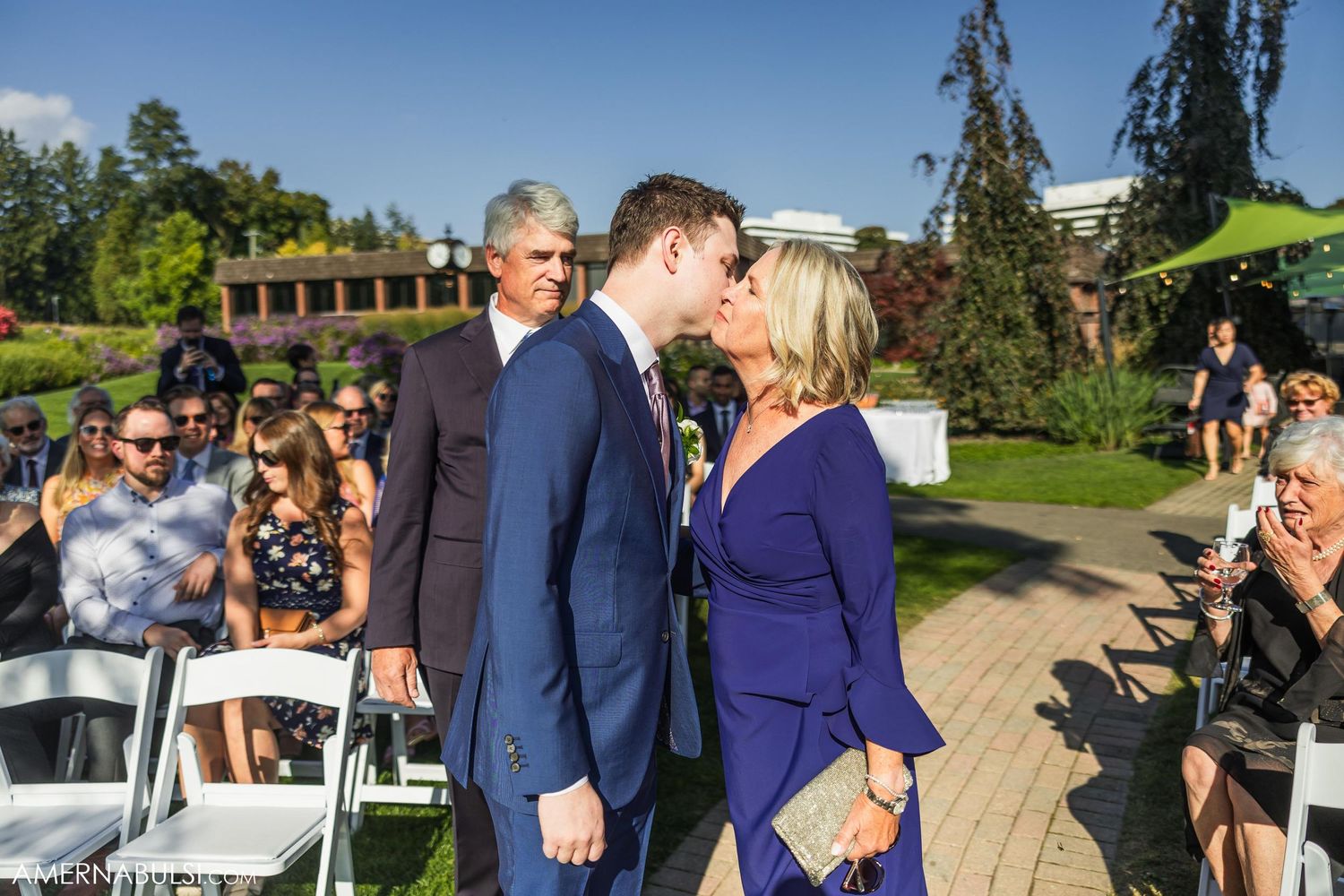 Outdoor wedding ceremony moment with guests seated on white chairs in a garden setting.
