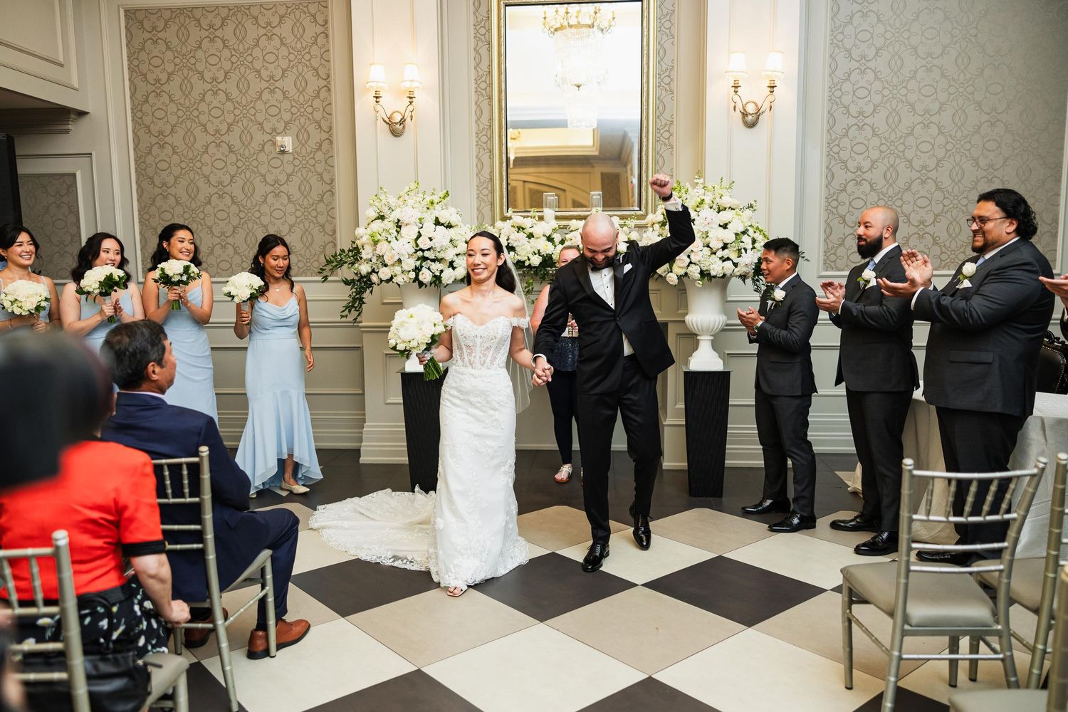 Wedding celebration in elegant ballroom with checkerboard floor and white floral decor.