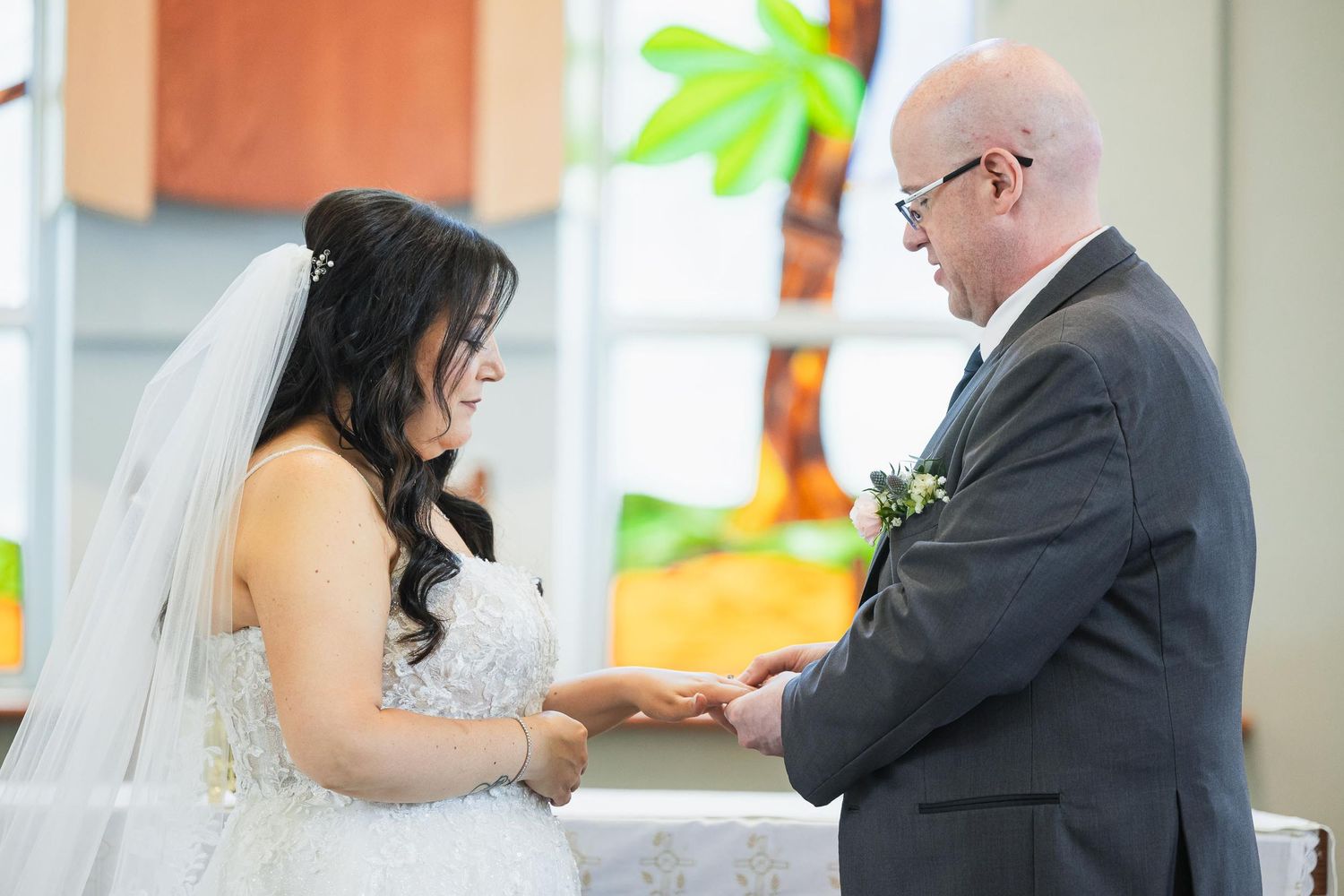 Ring exchange during wedding ceremony with colorful stained glass in background.