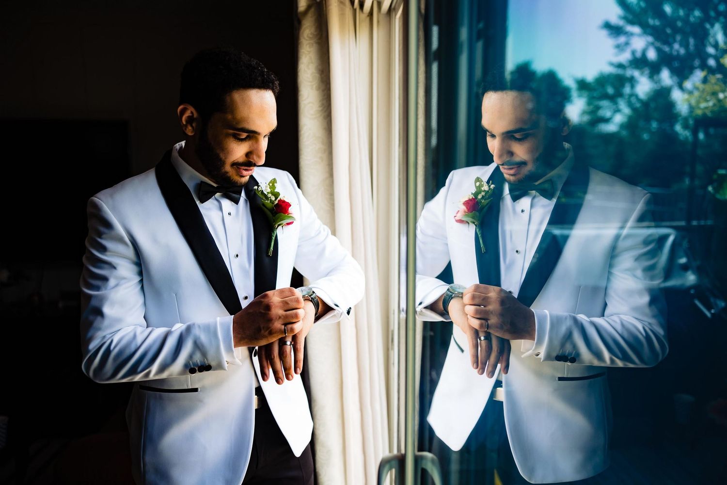 Groom in white tuxedo adjusts boutonniere while looking in mirror.