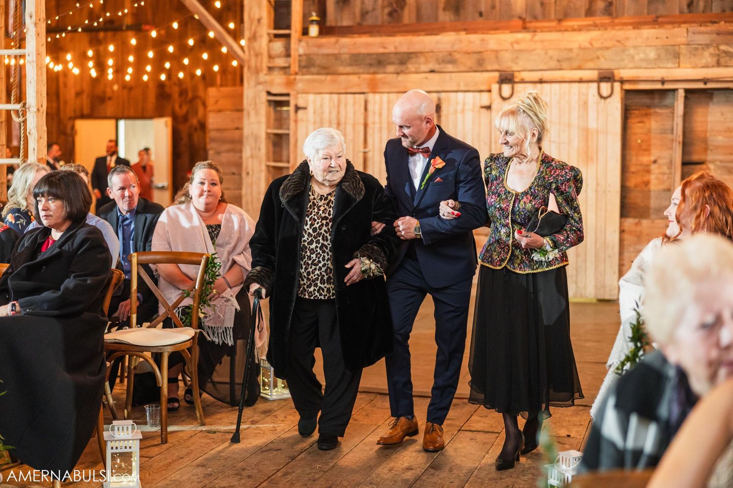 Wedding guests walking down aisle in rustic barn venue decorated with string lights and wooden details.